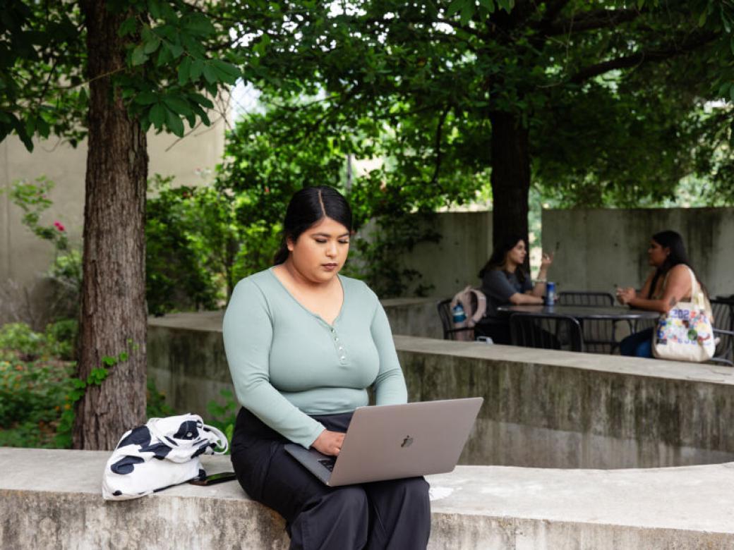 A student sits outside with a laptop on their lap and types. Trees and two other students sitting at a patio table are in the background.