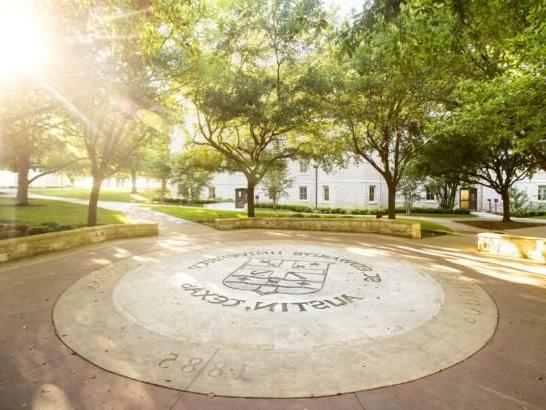 的 image depicts a paved circular area with the words "ST. EDWARD'S UNIVERSITY AUSTIN, TEXAS" and the year "1885" inscribed on it. 这个徽章或印章可能位于圣. 爱德华大学位于德克萨斯州奥斯汀的校园. 周围葱郁的绿树营造出一种宁静的气氛, 背景中可以看到带有多个窗户的白色建筑. 这幅图像传达了一种与大学有关的遗产和自然美景.